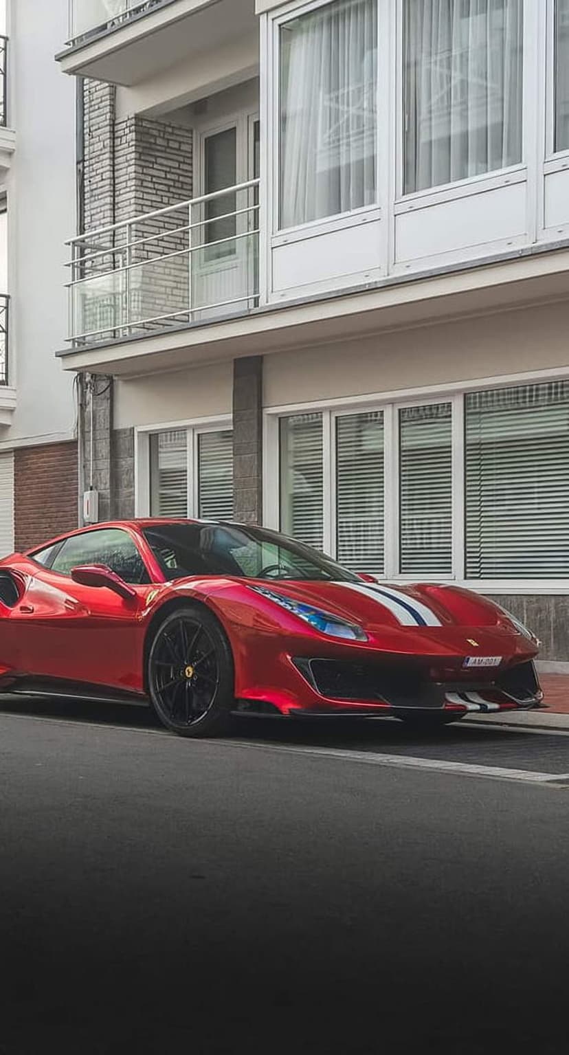 Red Ferrari supercar with white racing stripes parked on an asphalt street in front of a modern brick building.