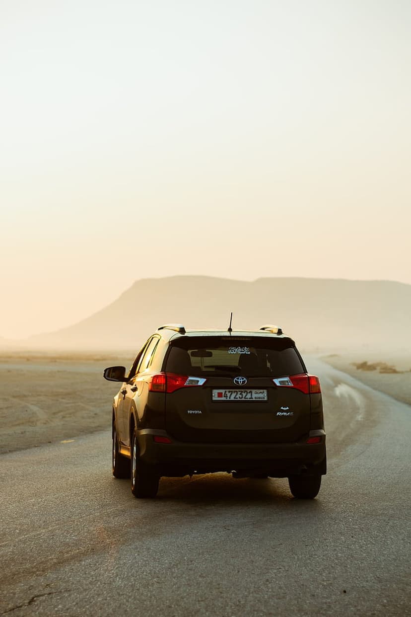 Dark SUV driving on a desert road towards hazy mountains under a warm golden hour sky during sunset.