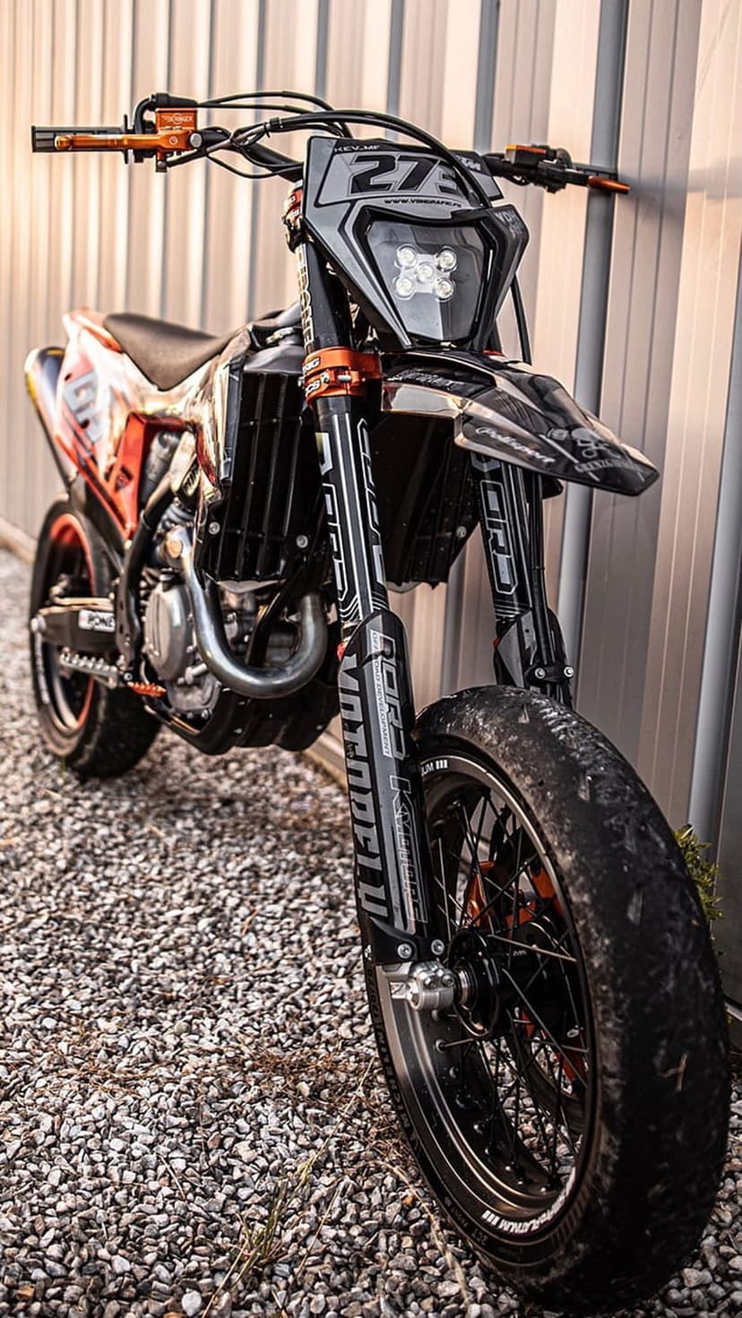 Orange dirt bike number 27 rests against a light metal wall on gravel during late afternoon golden hour.
