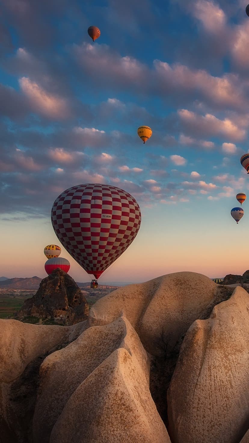 Checkered hot air balloons float above jagged rock spires and volcanic valleys during a soft pastel sunrise.