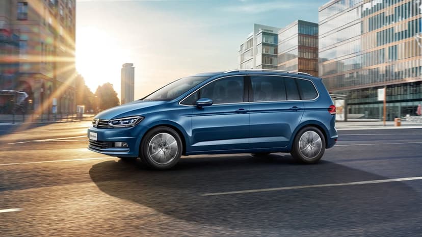Blue Volkswagen Touran driving on a sun-drenched city street with tall buildings and golden lens flare.