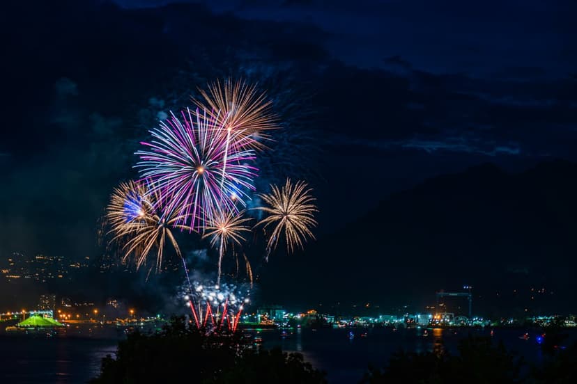 Brilliant pink and gold pyrotechnics burst above a dark urban skyline reflected in calm water at night.