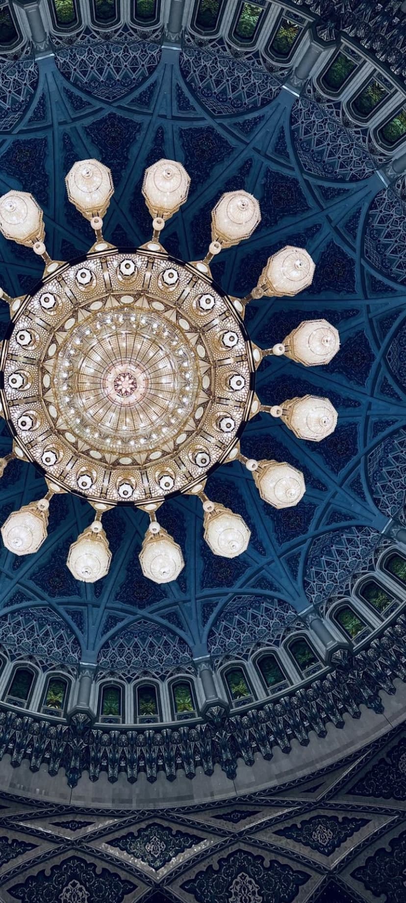 Golden chandelier hanging from a blue vaulted mosque dome with geometric patterns and arched windows.