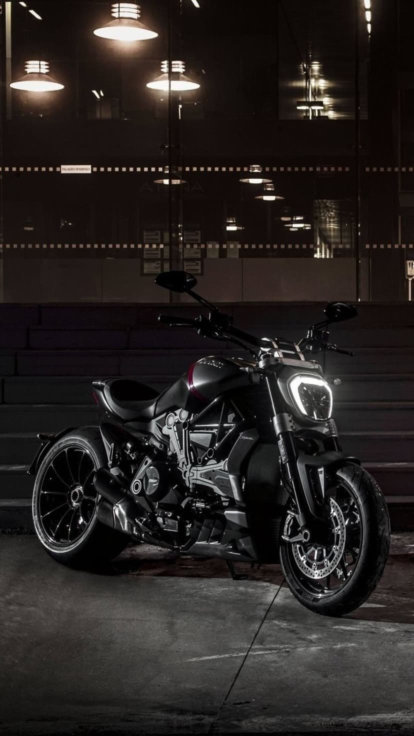 Black Ducati motorcycle with a glowing headlight parked on concrete near urban stairs under soft night lights.
