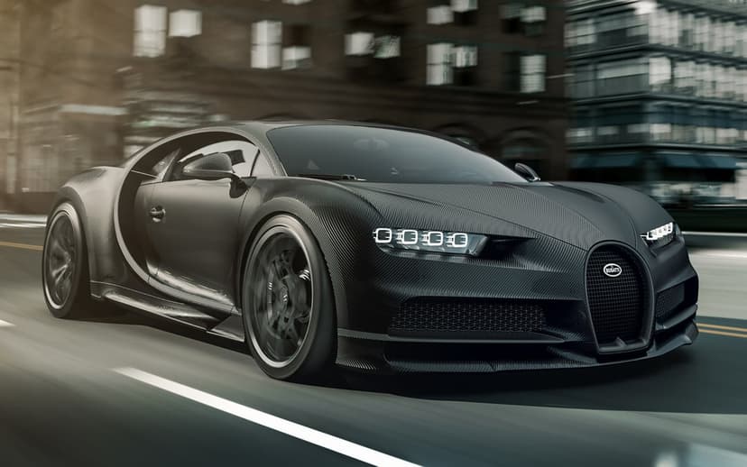 Matte black carbon fiber Bugatti Chiron hypercar driving fast on an urban road with motion blur and city buildings.