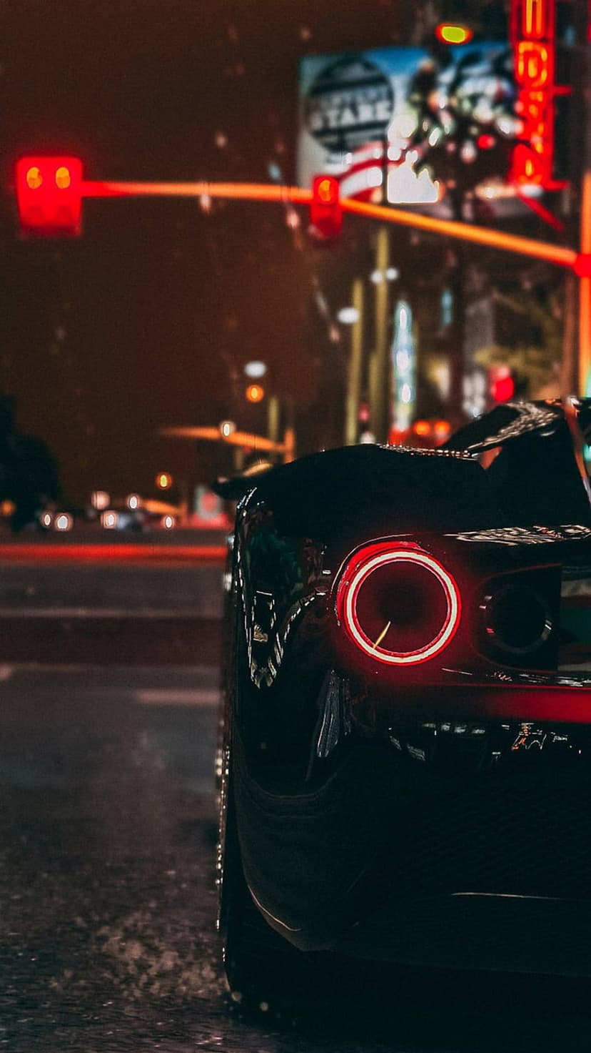 Red car taillights illuminate a wet asphalt road at night with colorful neon reflections and city lights.
