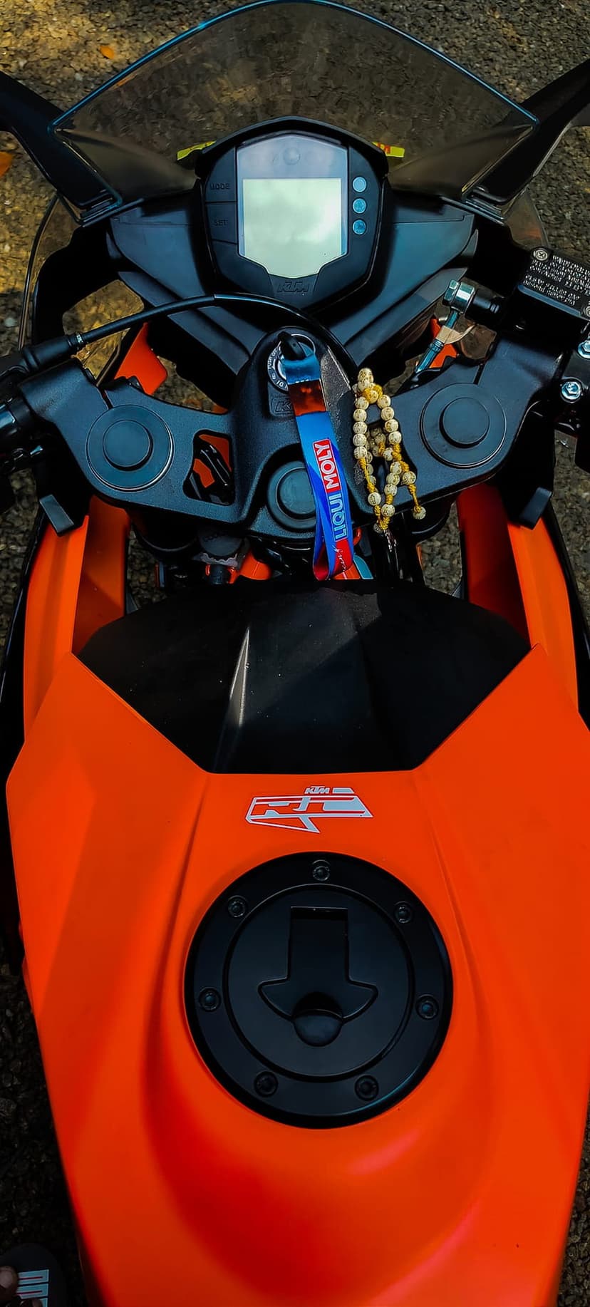 KTM RC motorcycle orange fuel tank and digital dashboard with a Liqui Moly keychain and hanging prayer beads.