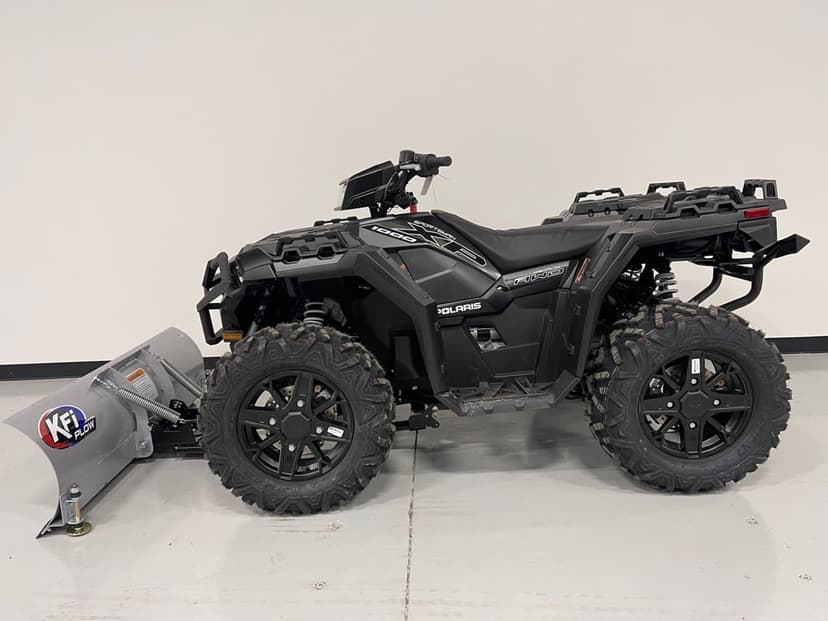 Black Polaris Sportsman XP 1000 ATV with a silver snow plow attachment on a grey floor against a white wall.