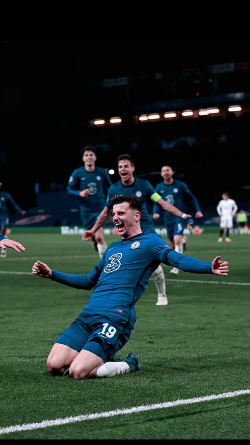 Mason Mount kneels with arms outstretched in a blue Chelsea kit while celebrating on a green football pitch.
