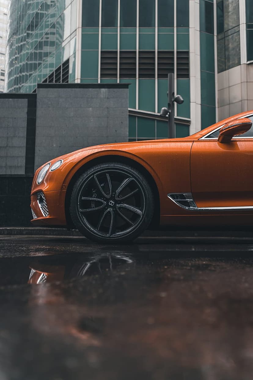 Orange Bentley Continental GT front wheel and fender reflecting on a wet city street during a rainy day.