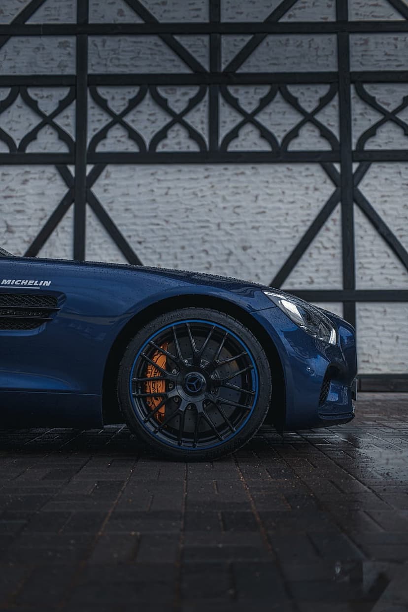 Mercedes-AMG GT front wheel with orange brake calipers on wet pavement next to a white brick wall.