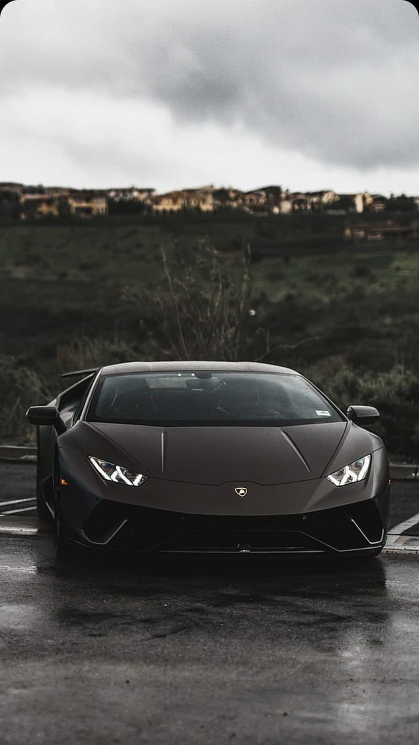 Dark Lamborghini supercar parked on wet asphalt with glowing headlights against a cloudy hill landscape.
