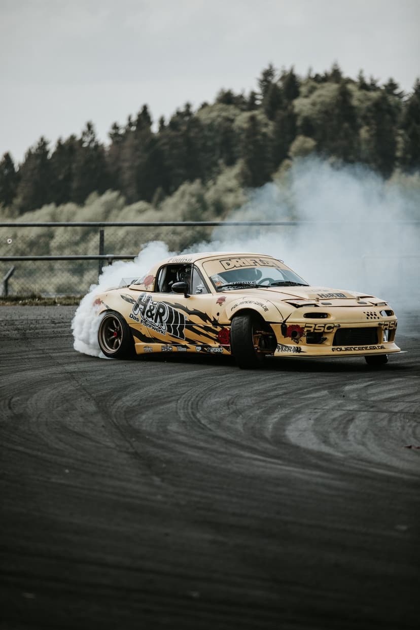 Yellow Mazda MX-5 Miata drifting on a race track with thick white tire smoke against a blurred forest backdrop.