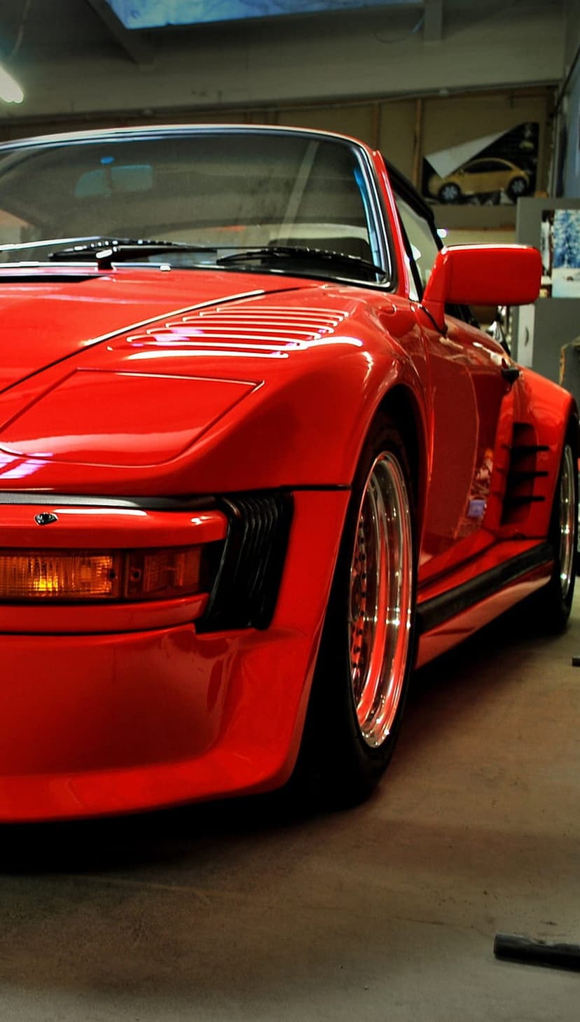Red Porsche 930 Turbo Slant Nose with a whale tail spoiler parked inside a workshop with reflective floors.