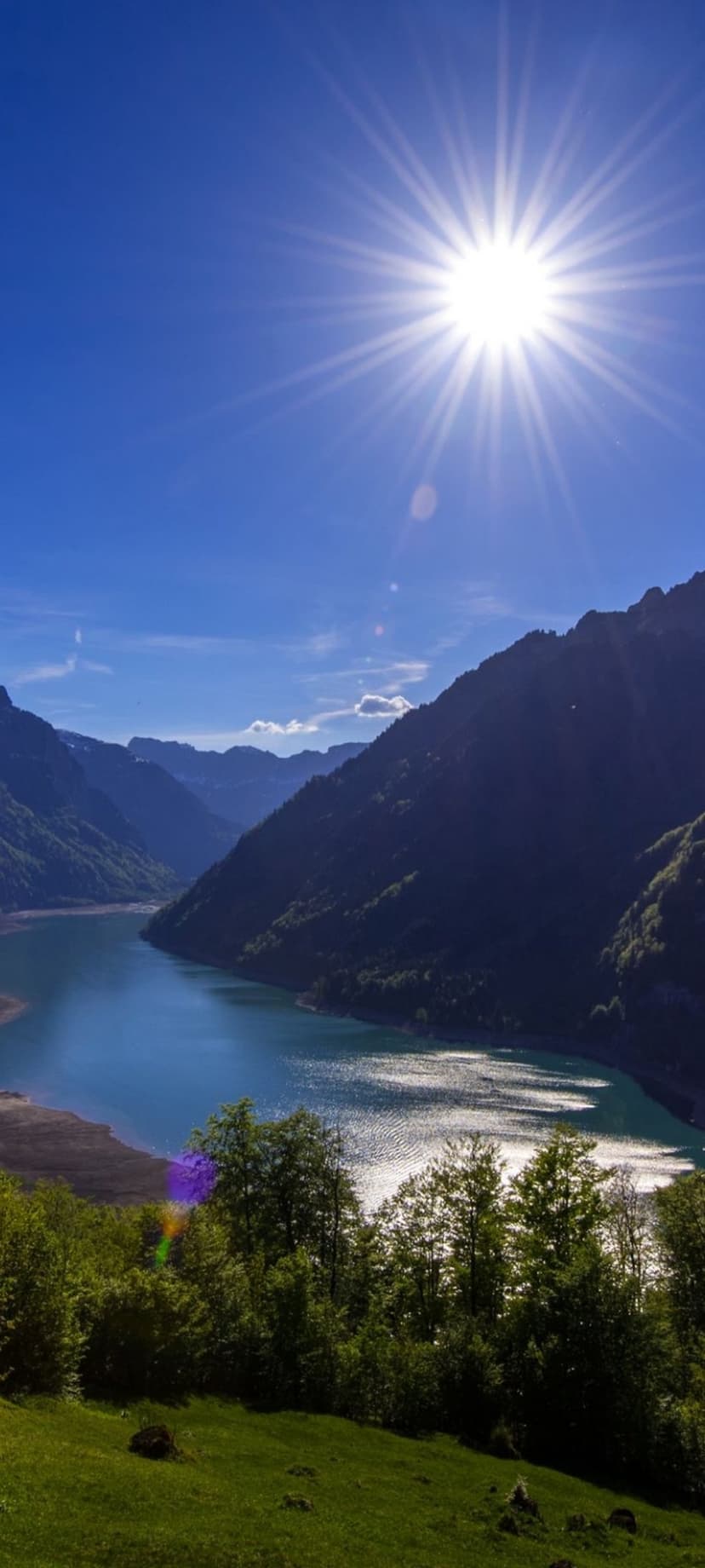Glacial alpine lake sits beneath sunlit granite peaks and a clear blue sky in a vertical mountain landscape.