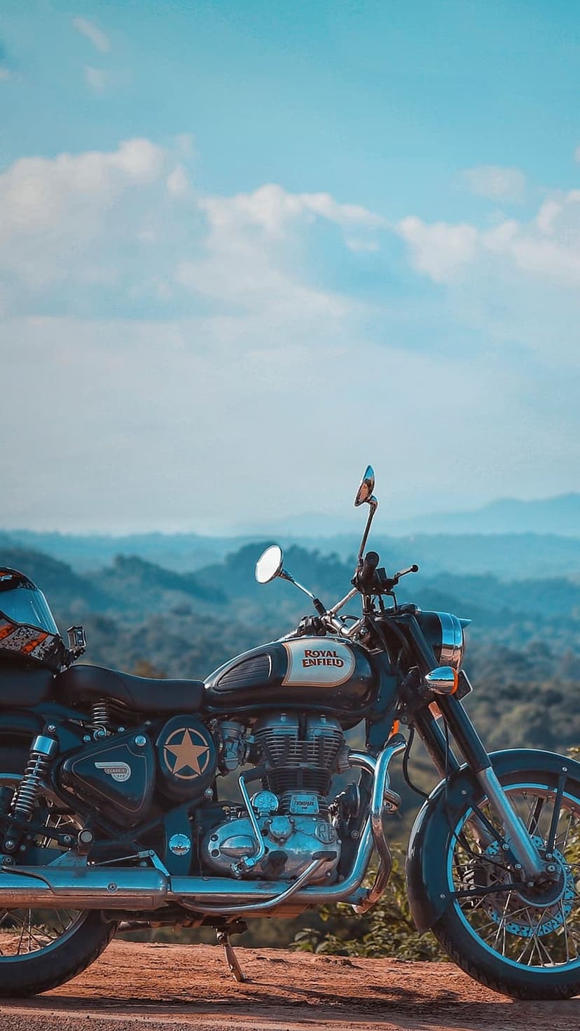 Royal Enfield motorcycle parked on a dirt trail overlooking lush green mountains under a bright cloudy sky.
