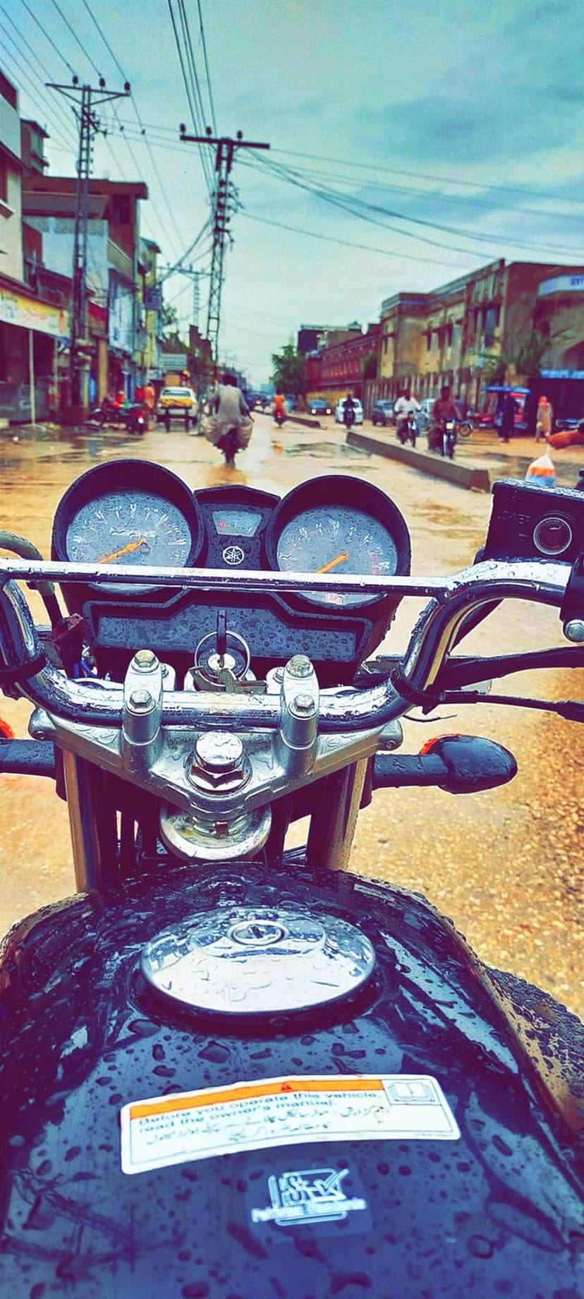Motorcycle fuel tank and dashboard covered in raindrops on a wet street with town buildings in the background.