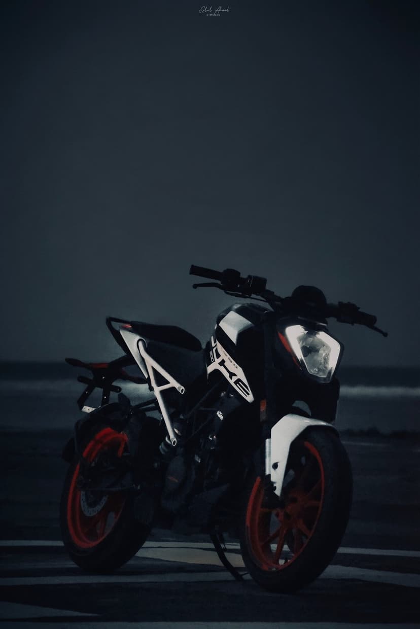 KTM Duke motorcycle with orange wheels parked on a dark sandy beach under a moody overcast sky at twilight.