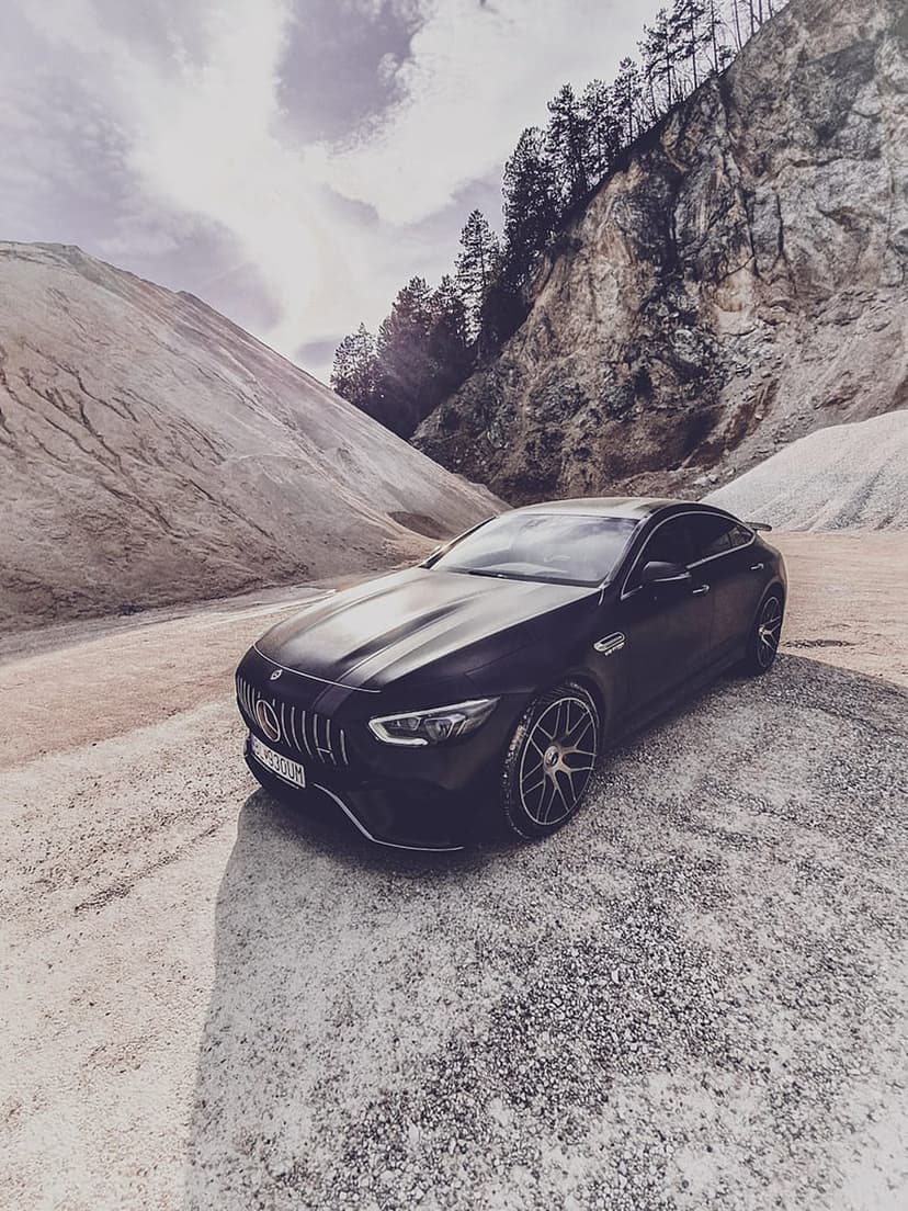 Black Mercedes-AMG GT 4-Door Coupe parked on gravel with steep rocky cliffs and a cloudy sky in the background.