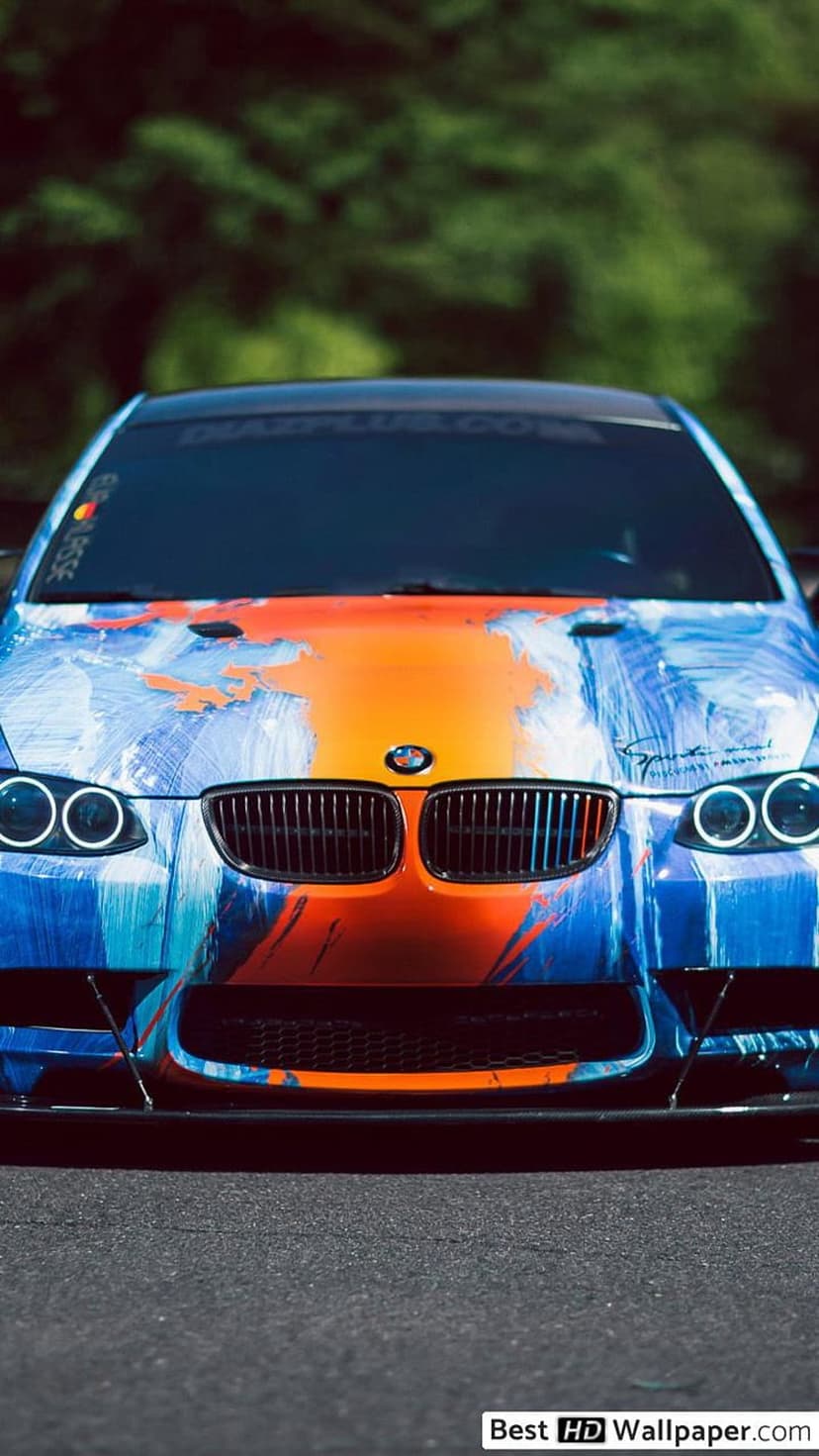 BMW M3 front view with blue and orange abstract paint splatters on an asphalt road with glowing headlights.