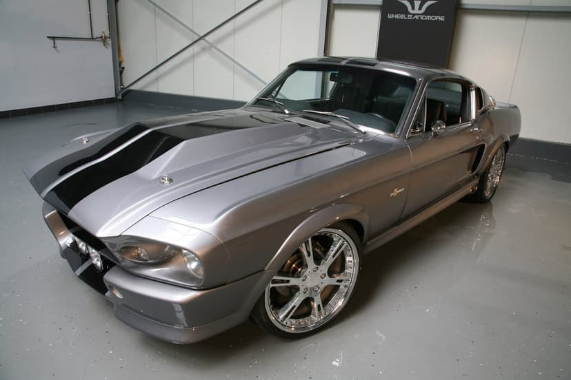 Silver 1967 Shelby GT500 Mustang with black stripes and chrome wheels parked in a modern grey showroom.