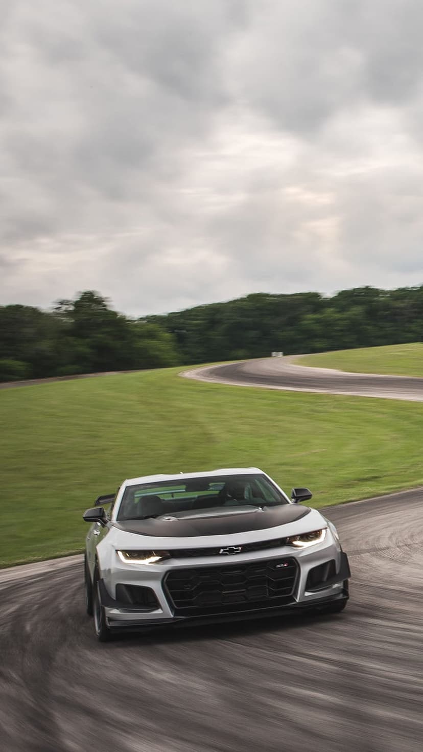 White Chevrolet Camaro ZL1 drifting on a wet racetrack with a black hood and overcast sky in the background.