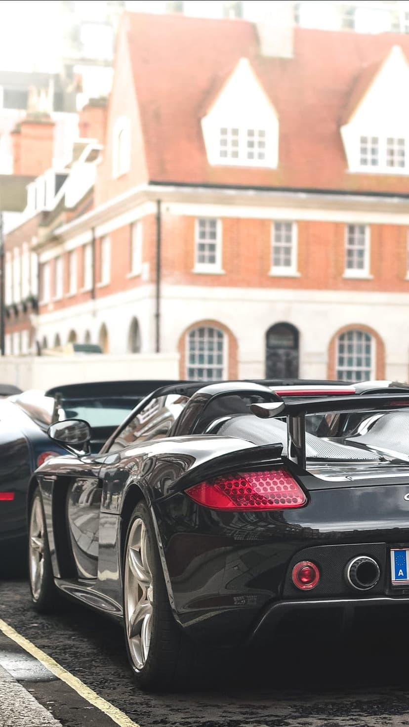 Black Porsche convertible with glowing red taillights parked on a European cobblestone street near brick buildings.