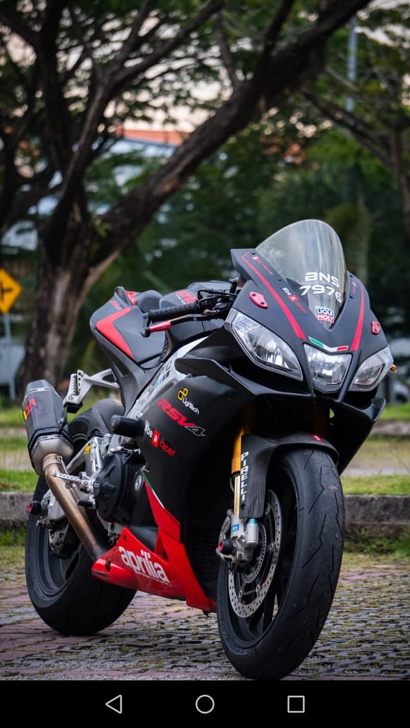 Aprilia RSV4 motorcycle in red and black livery parked on asphalt with a blurred tree background in daylight.