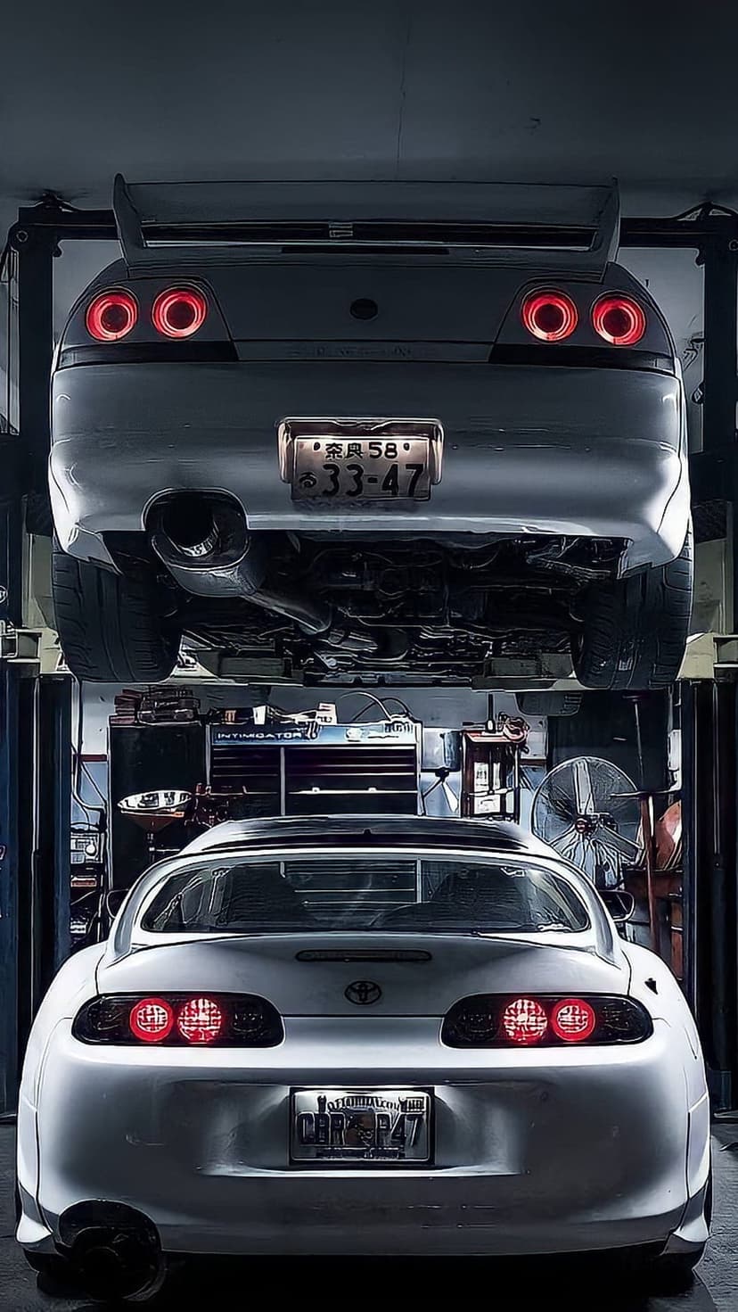 Two white Toyota Supras parked in a dark garage with one car raised on a lift showing its rear exhaust pipes.