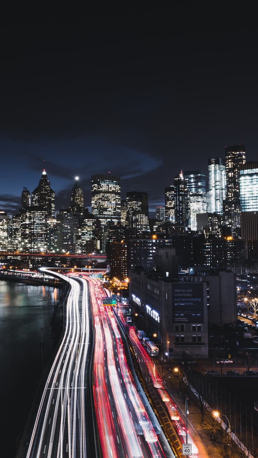 Glowing traffic light trails lead toward illuminated skyscrapers under a dark night sky in a vertical cityscape.