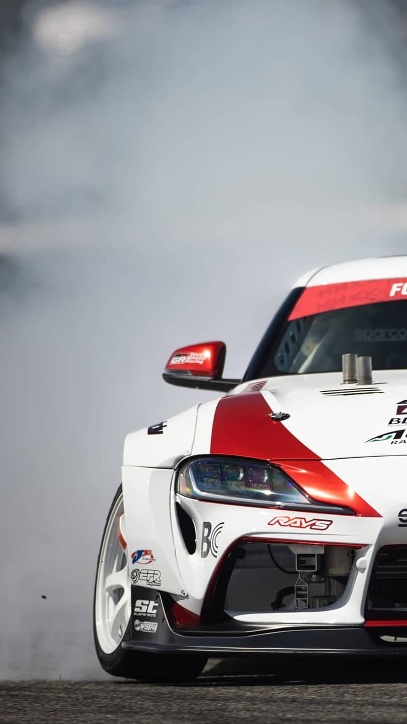 Toyota Supra in a red and white racing livery drifts while creating a thick cloud of white smoke on a track.