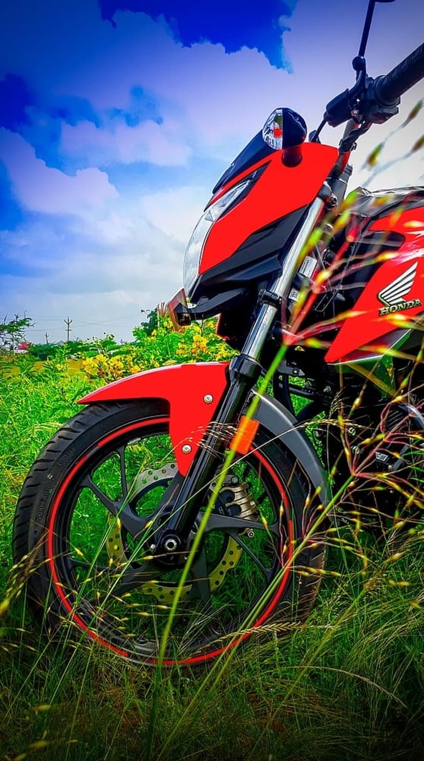 Red Honda motorcycle with front wheel detail parked in tall green grass under a dramatic blue sky with white clouds.