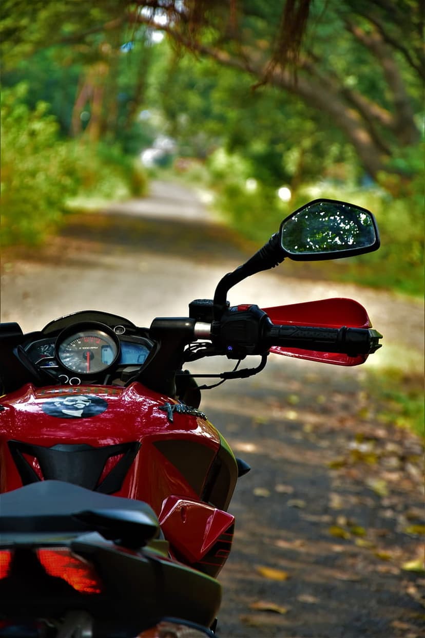 Red motorcycle handlebar and rearview mirror reflecting a dirt road surrounded by dense green forest trees.