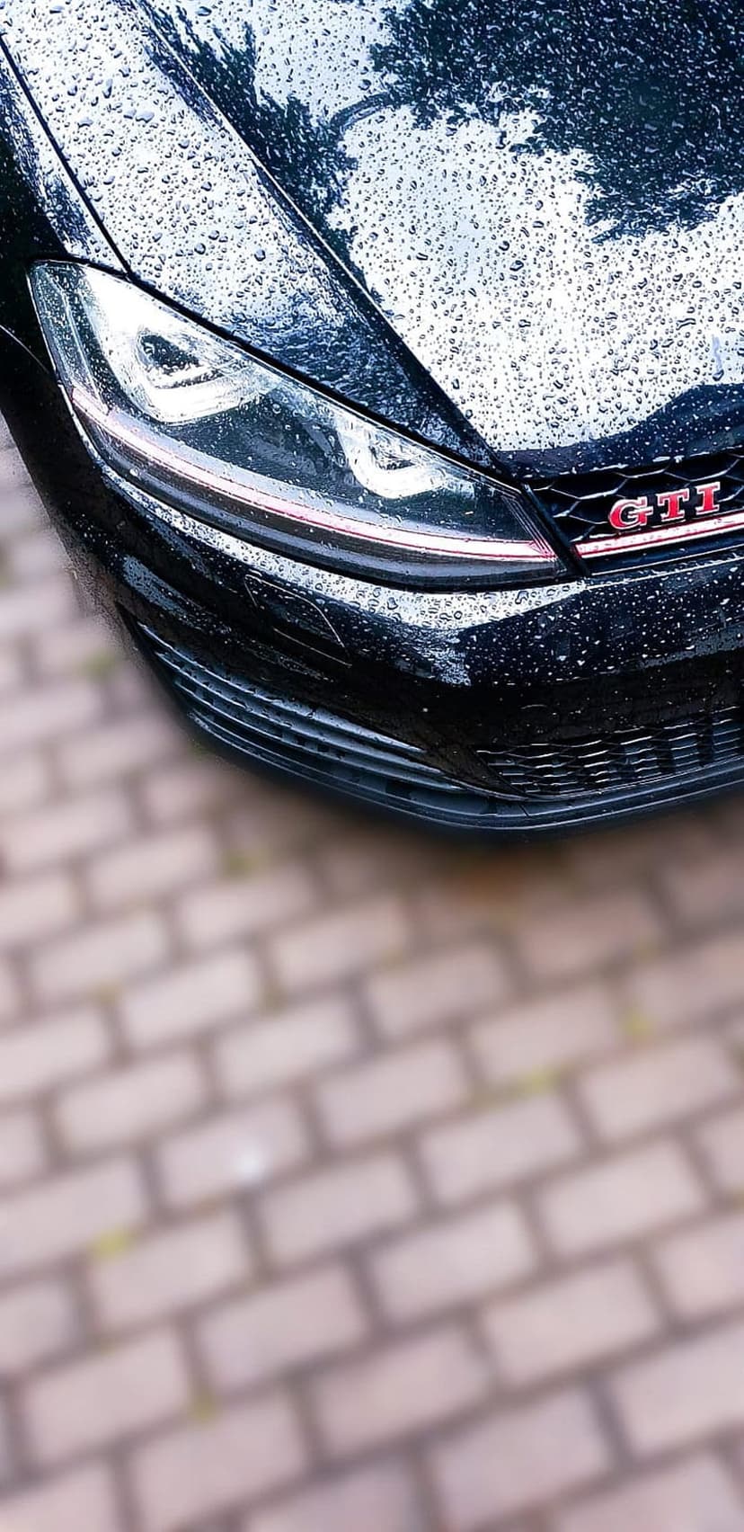 Red GTI badge and glowing LED headlight on a wet black Volkswagen Golf parked on blurred cobblestone pavement.