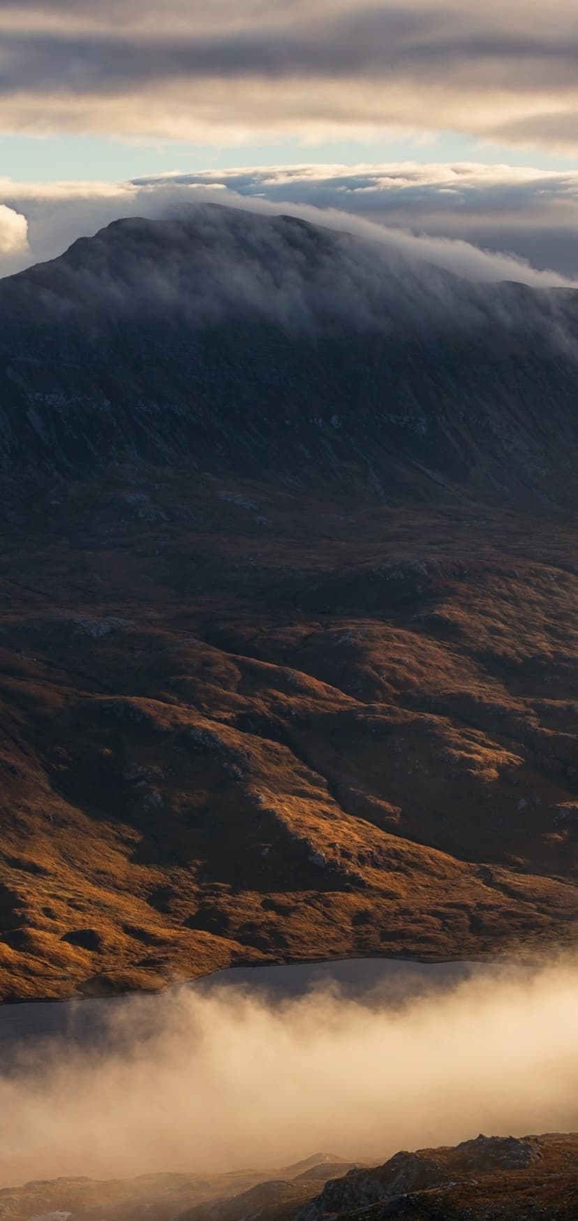 Foggy mountain peaks rise above a still lake at sunrise with orange light filtering through thick morning mist.