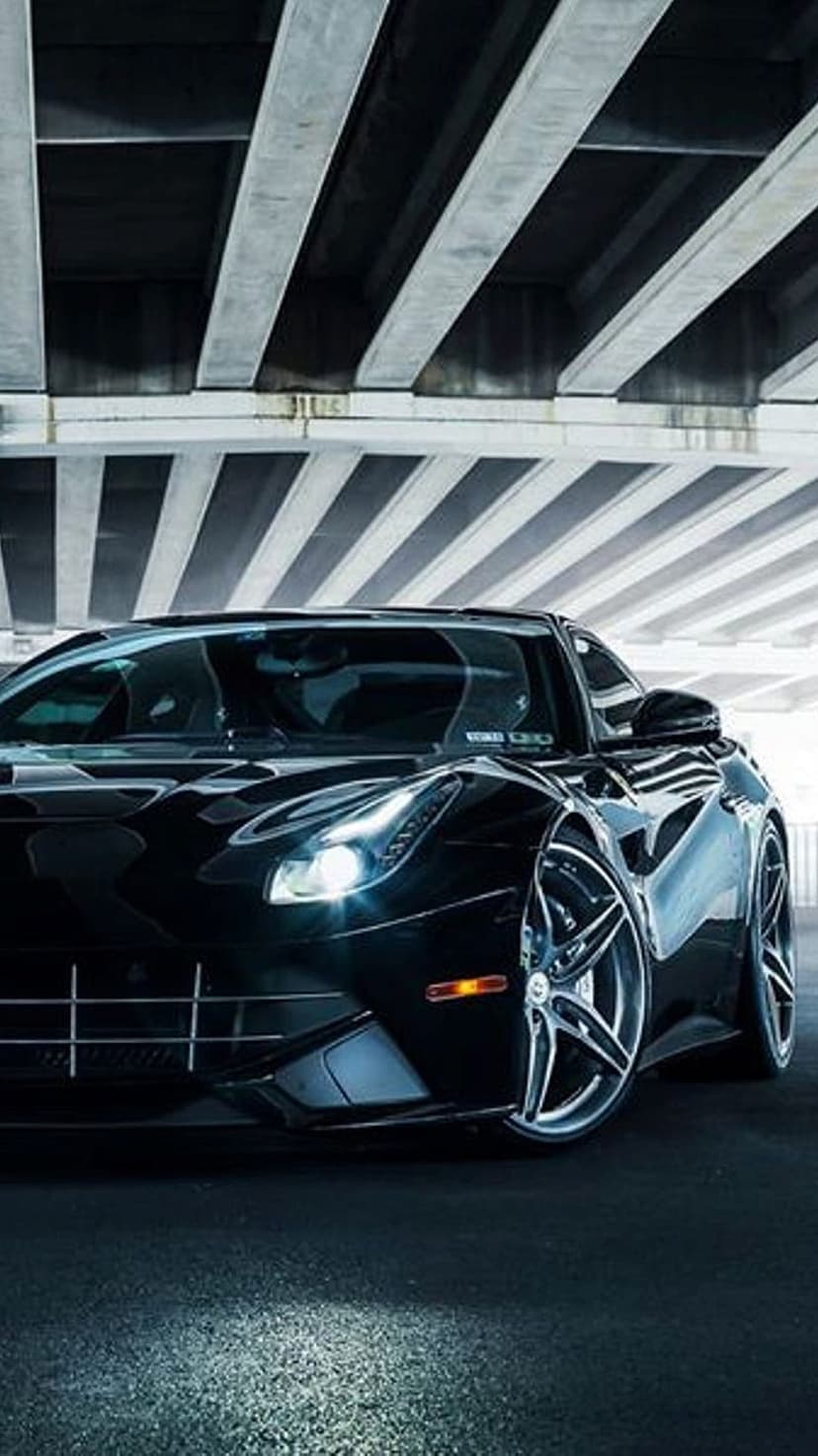 Black Ferrari supercar under a concrete bridge with harsh lighting on the front grille and alloy wheels.