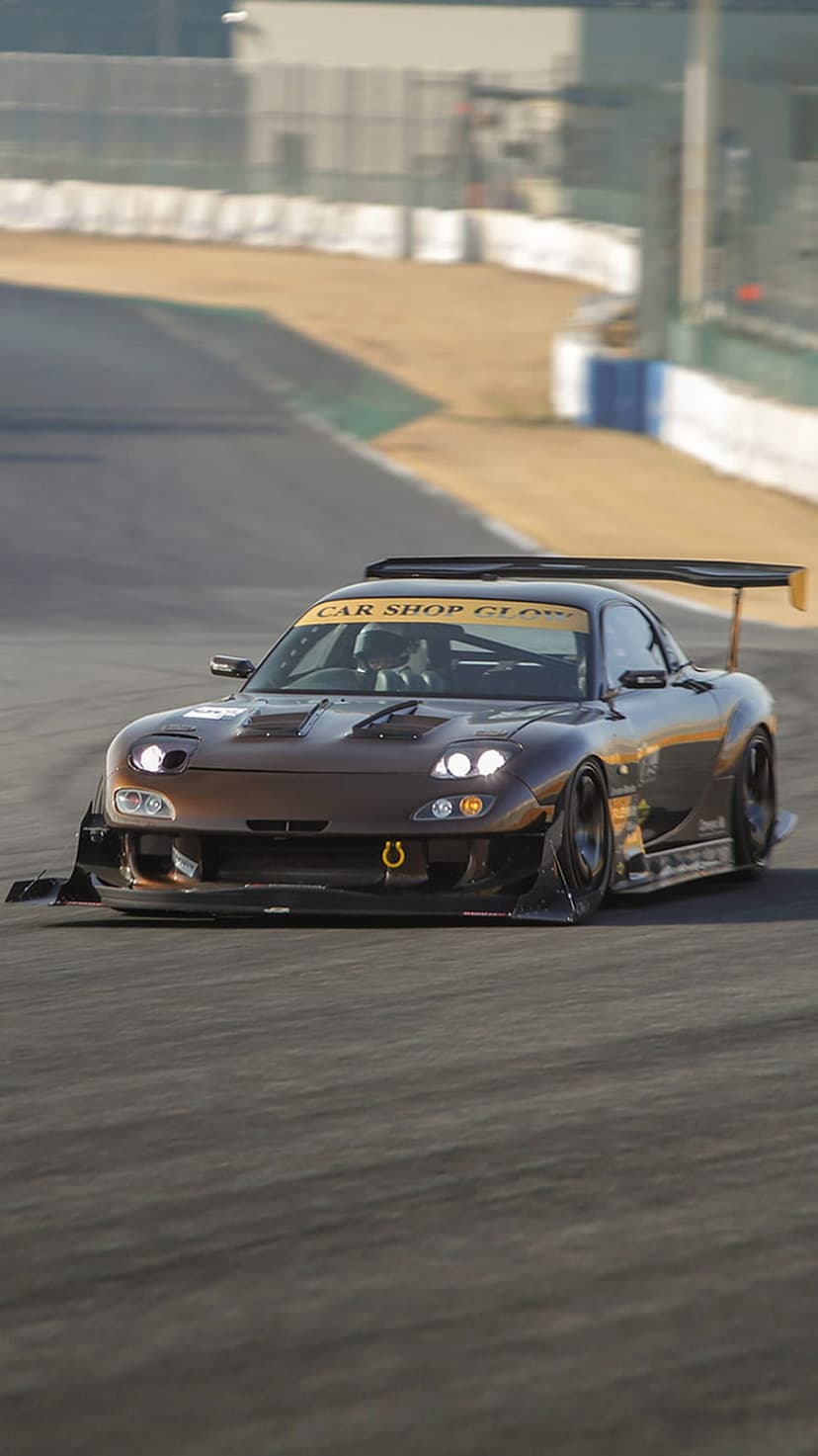 Brown Mazda RX-7 FD with a widebody kit and rear wing drifting on a race track with smoke and motion blur.