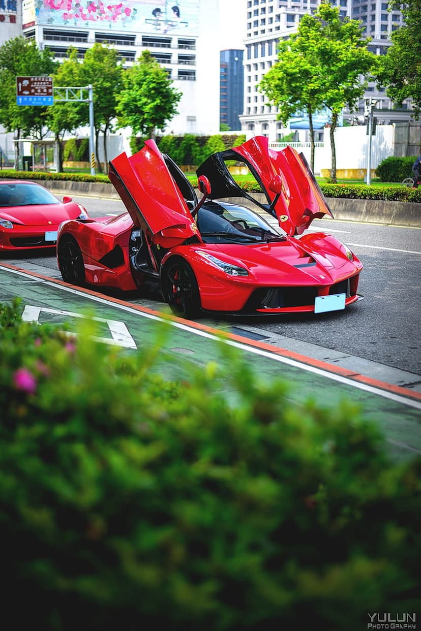 Red Ferrari supercar with open scissor doors parked on a paved city street with modern buildings in the background.