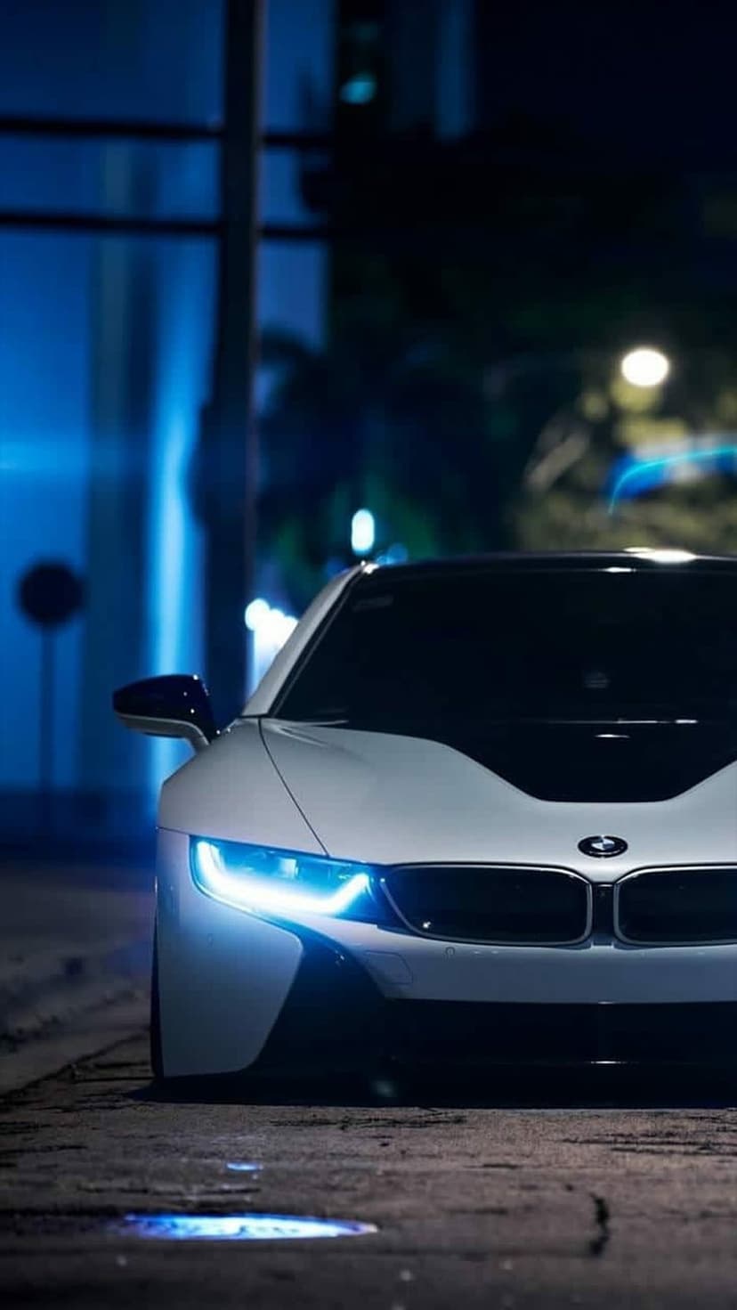 BMW i8 front headlights glow blue on a dark road with blurred urban lights in the background at night.