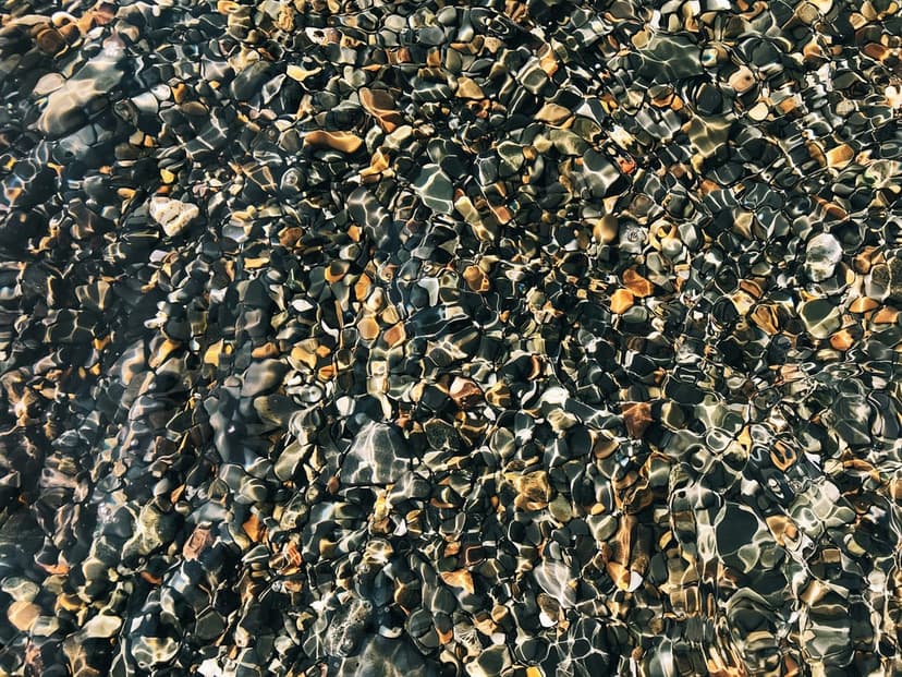 Smooth multi-colored pebbles lie underwater with light refracting through rippled waves in a top-down view.