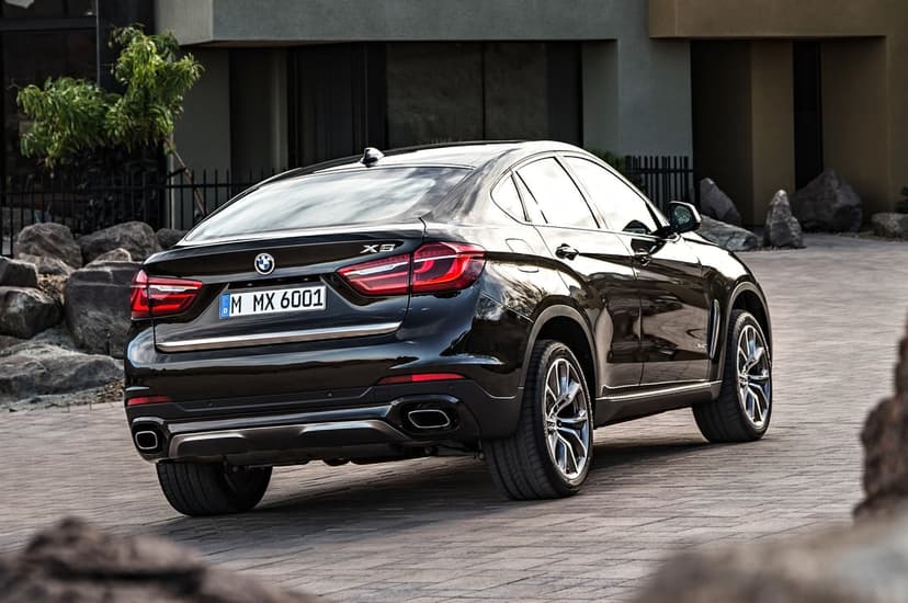 Rear view of a black BMW X6 parked on a paved driveway in front of a modern house with rocks and a small tree.