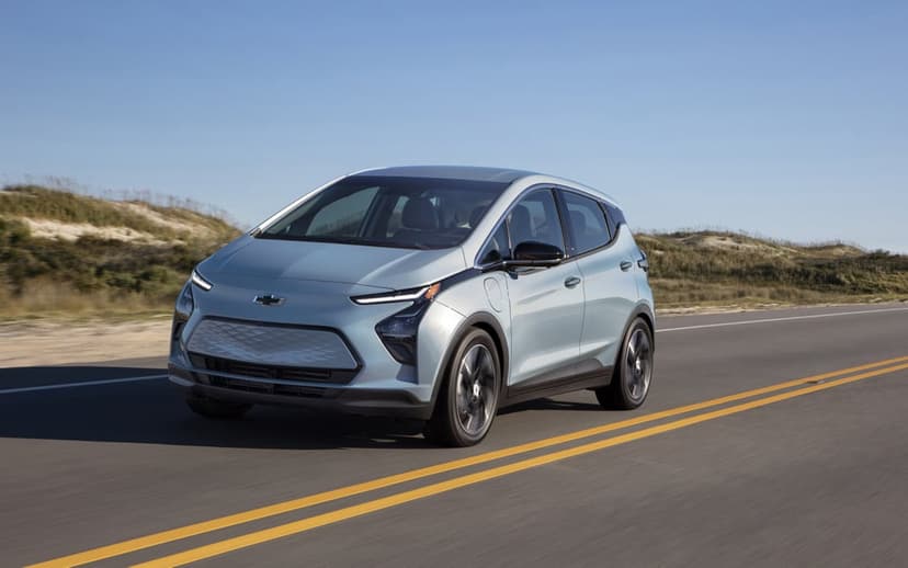 Light blue Chevrolet Bolt EV driving on a paved coastal road beside sand dunes under a clear blue sky.
