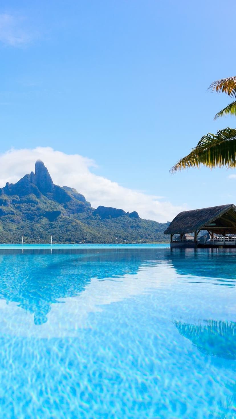 Turquoise infinity pool borders a blue lagoon with a thatched hut and green mountains under a bright clear sky.