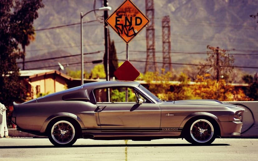 Silver Shelby GT500 muscle car in profile on a road with an END traffic sign and mountains in the background.