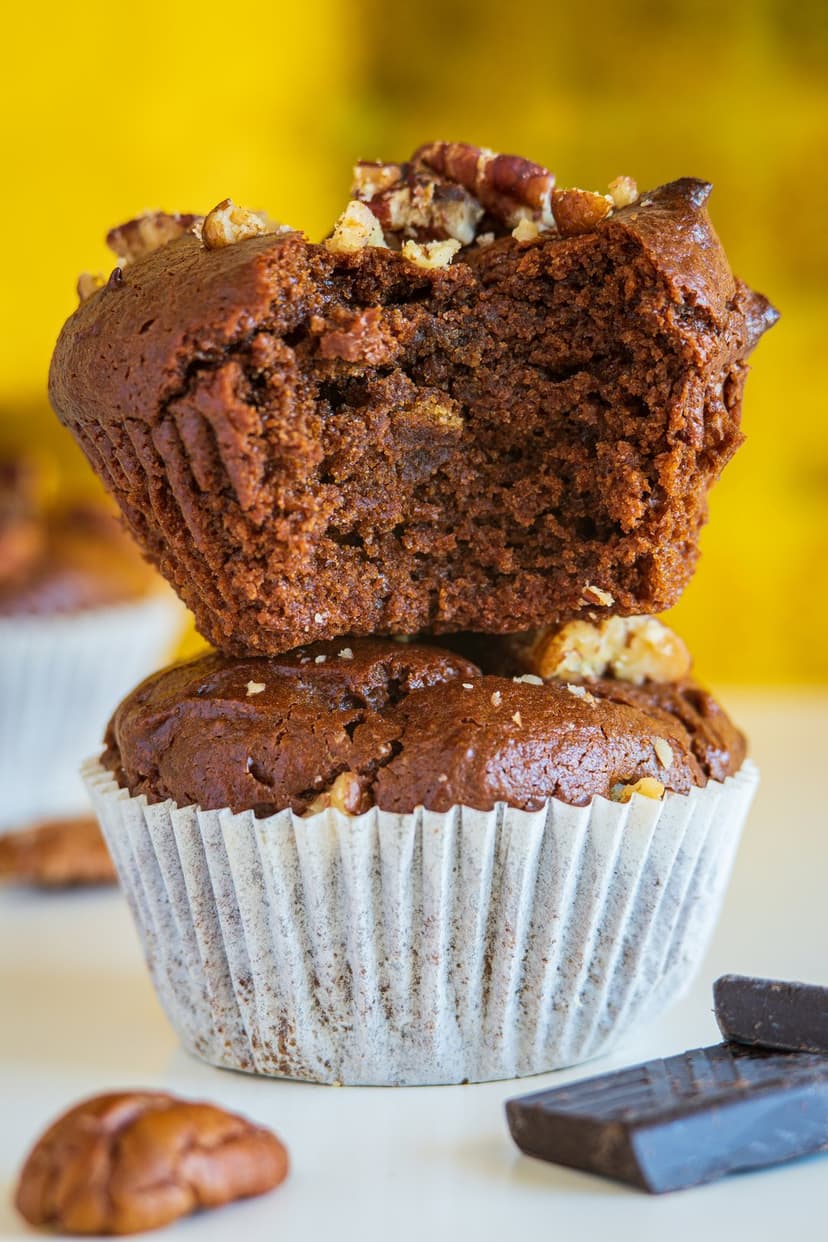 Stacked dark chocolate muffins topped with crunchy pecans and nuts against a soft-focus bakery background.