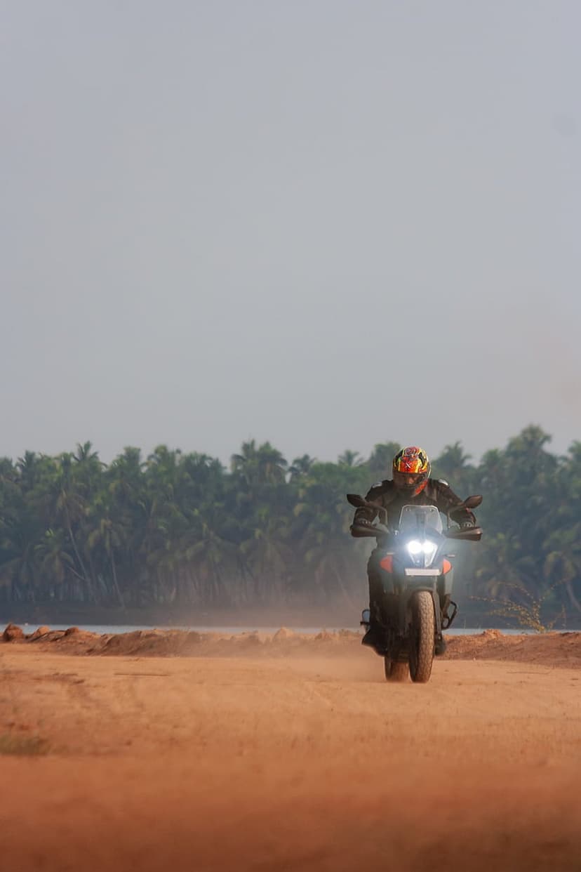 Motorcyclist in full gear rides an adventure bike on a dusty beach path lined with tall palm trees and a hazy sky.