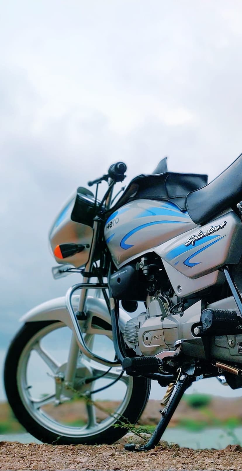 Silver and blue Hero Splendor motorcycle showing engine and wheel details against a cloudy outdoor sky.