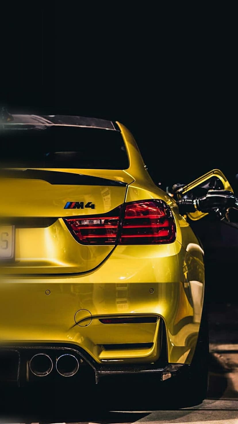 BMW M4 in bright yellow parked at a gas station at night showing rear taillights and quad exhaust pipes.
