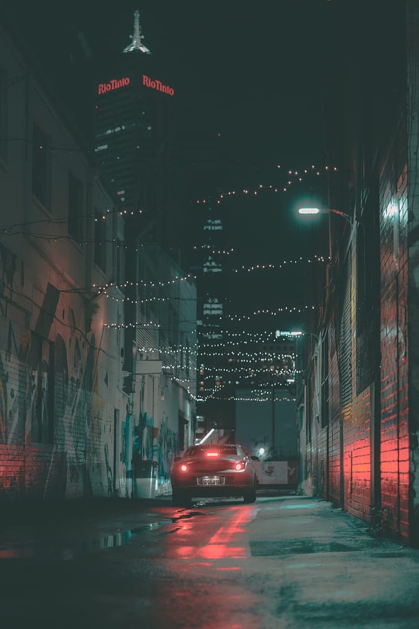 Red car taillights reflect on wet pavement in a dark alley with graffiti and a distant lit skyscraper.