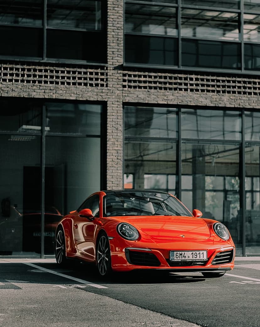 Orange Porsche 911 parked in front of a modern brick building with large glass windows and black roofing.